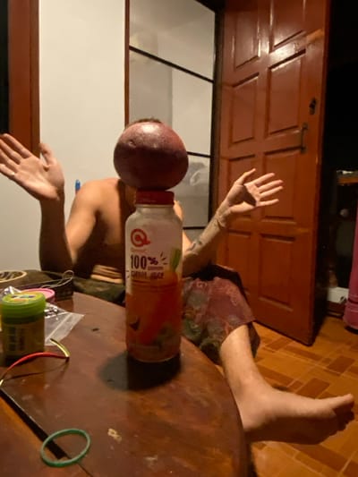 This image depicts an indoor scene in Haad Yao, Thailand. A dark brown wooden table occupies the foreground. Centrally positioned on the table, a dark red, spherical fruit is balanced atop the red cap of a white plastic bottle. The bottle's label displays "Q Omin C 100% CARROT JUICE." To the left of the bottle on the table, a green-capped container, a small transparent plastic bag containing small components, two thin wires (one red, one gold), and a green rubber band are visible. A shirtless individual is seated behind the table, largely obscured by the central objects. Their face is not visible. Both of their hands are raised and open, positioned to the left and right of the balanced items. The individual's right leg, partially covered by patterned fabric, extends onto a light brown tiled floor. In the background, a reddish-brown wooden door is ajar on the right, revealing a dark exterior and a pink object on the tiled floor just beyond the doorway. To the left of the door, a white wall with a dark vertical panel is visible. The scene is illuminated by artificial light, casting shadows from the bottle onto the table.