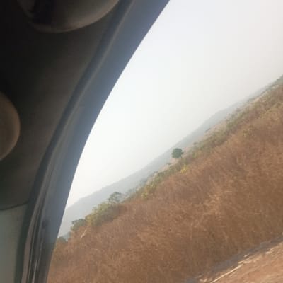 The image depicts an exterior landscape viewed obliquely from inside a vehicle, located in Assob Hausa, Nigeria. The left side of the frame is occupied by dark gray vehicle interior components, including a curved pillar and a partial view of a lighter, beige circular object, likely a handle or light fixture, in the upper left. The exterior scene, seen through a window, shows a sloping, undulating terrain covered predominantly in dry, light-brown vegetation, consistent with dry grasses or shrubs. Scattered green trees and bushes are visible, particularly along the mid-slope and in the distance; one solitary green tree is prominent in the mid-upper portion of the landscape. A faint, hazy ridge line is discernible in the far background. The sky is bright and uniformly pale, suggesting atmospheric haze or strong daylight. A light-brown, unpaved surface, possibly a dirt road or path, is visible at the bottom right. No individuals or specific actions are depicted in the exterior environment.