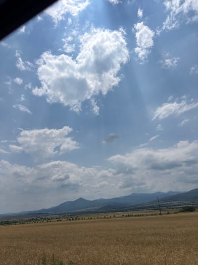 The image depicts a landscape with a field of golden grain in the foreground. Beyond the field, a line of rolling hills and mountains stretches across the horizon under a partly cloudy blue sky. A single, tall, dark pole stands in the middle-right section of the frame, possibly a utility pole. The sky is illuminated by shafts of sunlight breaking through the clouds. The scene appears to be in a rural area.