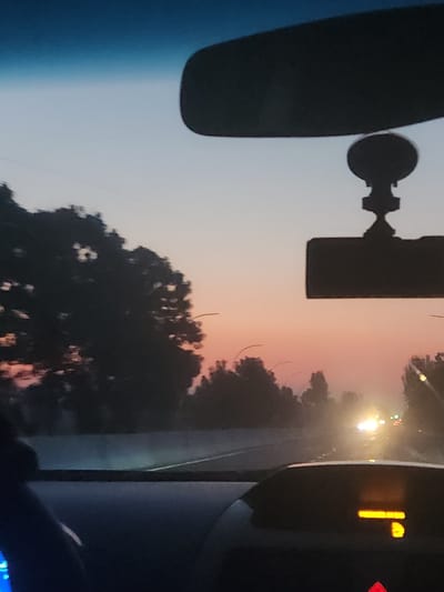 The image depicts a view from inside a vehicle at dusk, with the sky exhibiting hues of blue, pink, and orange. The dashboard of the car is visible in the foreground, featuring an illuminated orange digital display and a red warning light. A rearview mirror is mounted on the windshield, obscuring a portion of the sky. Outside the vehicle, silhouetted trees line the road, and distant headlights of other vehicles are visible through a hazy atmosphere. The scene is consistent with a drive on a highway outside the city of Kipchak, Uzbekistan, during twilight.