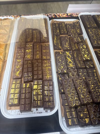 This media file shows an overhead view of several trays filled with artisanal chocolate bars. The chocolates are dark brown and have decorative gold accents on their surfaces. Some bars are divided into smaller squares, while others feature intricate geometric patterns, possibly starbursts or faceted designs. The chocolates are arranged neatly in white trays, with some placed on brown paper liners. The trays are stacked or placed next to each other, suggesting a display in a shop or market. In the background, there's a glimpse of a printed design on what appears to be packaging or a display element. The lighting suggests an indoor setting, possibly a bakery, confectionery shop, or a market stall in Ocean City, USA. No people are visible in the frame, and the focus is entirely on the confectionery items. The image is well-lit, highlighting the textures and details of the chocolates. There is no visible text in the frame.