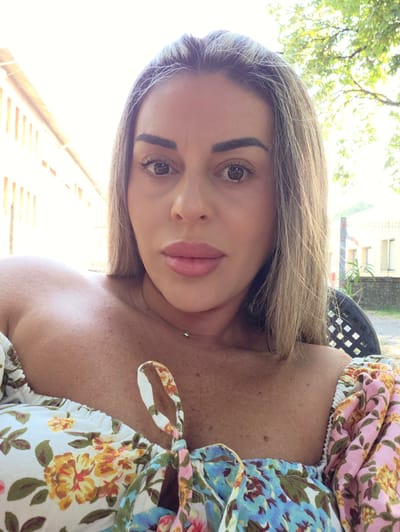 A medium shot, eye-level outdoor selfie shows a woman with blonde hair and fair skin. She is wearing a floral off-the-shoulder top in shades of white, pink, yellow, and blue. The woman is looking directly at the camera. Her makeup includes eyeliner, mascara, and light pink lipstick. She has a light-colored necklace with a silver charm around her neck. Her skin has some freckles and small bumps. The background shows a building with a yellow facade and orange-striped awnings on the left, and trees and a building with windows on the right. A black chair with a diamond pattern is visible on the right side of the image. The lighting suggests it is daytime. No specific location in Correggio, Italy, can be identified from the visual information.