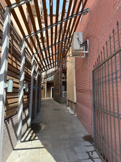 The image shows an outdoor alleyway with a tiled ground surface and brick walls on either side. A wooden slatted overhang with metal supports runs along the alleyway, casting shadows on the ground. Several outdoor light fixtures are attached to the wooden wall on the left. On the right, a rusted metal fence with decorative circular elements runs along the brick wall. An outdoor air conditioning unit is mounted on the brick wall. The scene appears to be in an urban environment, possibly in a commercial or residential area. There are no people visible in the image. The sky is visible at the top of the frame, suggesting it is daytime.