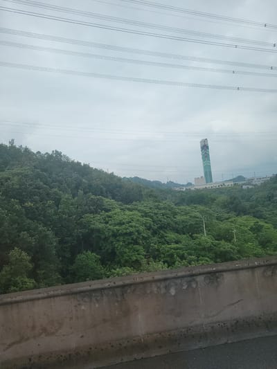 The image depicts a scenic view from what appears to be a highway or elevated walkway overlooking a densely wooded hillside. A prominent feature is a tall, slender building with a green and white facade, adorned with red Chinese characters. This structure stands out against the lush green of the trees and the hazy sky. In the foreground, a concrete barrier or wall runs along the edge of the pathway, with an asphalt surface visible on the right side. Overhead, a network of power lines stretches across the frame, suggesting the presence of infrastructure in the area. The overall impression is one of a landscape that blends natural beauty with urban development. The time of day appears to be daytime, with overcast or cloudy weather conditions, indicated by the diffused light and the soft, muted colors. The location context provided, Yufengshan, China, aligns with the visual cues of a hilly, vegetated landscape that might be found in such a region, possibly near an industrial or developed area given the power lines and the distinct building. The image does not convey any specific emotions, but rather a sense of observation or passage through a varied environment.