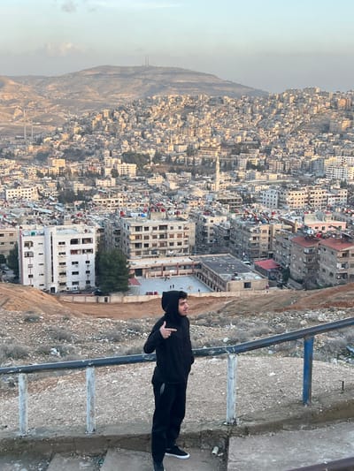 The image displays an expansive urban landscape identified as the city of Qudsaiya, Syria, observed from an elevated, barren hillside. In the foreground, a single male individual stands on a concrete surface, partially obscured by a metal railing with blue and white sections. He is dressed in a black hooded sweatshirt with the hood up, black trousers, and dark footwear. His right arm is bent, with his index finger extended, pointing towards the right side of the frame. His head is turned to the right, away from the central cityscape.

The midground and background reveal a densely populated area characterized by numerous multi-story residential buildings, primarily constructed from light-toned concrete or stone. These structures are closely packed and extend across rolling hillsides. A tall, slender white minaret with a pointed top is visible in the midground, rising above the surrounding buildings. Below and to the left of the foreground subject, a large building complex featuring a flat roof and an open, paved courtyard is discernible, possibly a school, with several small figures visible within the courtyard. Many building rooftops are equipped with water tanks or solar heating units. The distant background comprises arid, rocky mountains, with several communication towers or similar structures visible on the highest peaks. The immediate foreground ground is rough, stony, and sparsely covered with dry vegetation and scattered debris. The sky is a pale blue with minimal cloud cover, indicating daytime conditions.