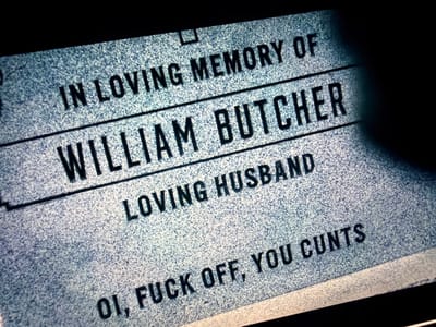 The image shows a close-up of a headstone. The inscription reads: "IN LOVING MEMORY OF WILLIAM BUTCHER, LOVING HUSBAND, OI, FUCK OFF, YOU CUNTS." The headstone appears to be made of light-colored stone with a grainy texture. Part of a dark object obscures a portion of the upper right corner of the image. There is no indication of location.