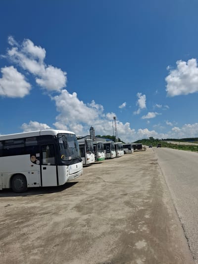 A fleet of white buses is parked on a gravel lot next to a paved road. The buses are lined up in a row, facing towards the right side of the image. The sky is blue with scattered white clouds. In the background, there are some buildings and trees. The scene appears to be in an industrial or rural area.
