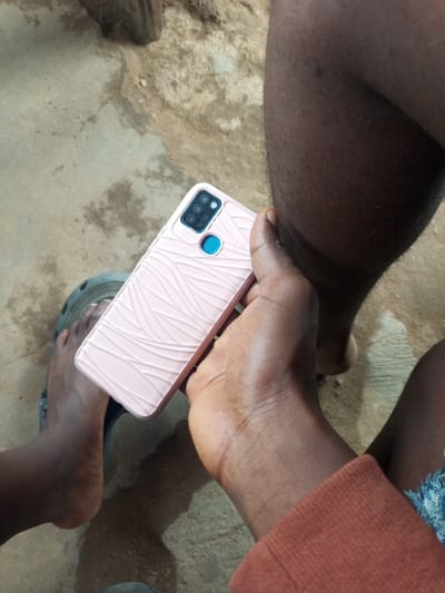 In Iyara, Nigeria, an individual's hand holds a pink mobile phone with a textured back. The phone is positioned in the foreground, partially obscuring the person's bare feet and a portion of their legs. The environment appears to be outdoors, with a concrete or dirt ground visible beneath. The image is taken from a high angle, looking down at the subjects and the phone.
