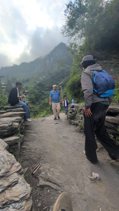 The image depicts a scenic mountain path in Malana, India, with several individuals present. In the foreground, a person wearing a grey jacket and a blue backpack is walking away from the camera. To the left, two young men are seated on large stones, with one wearing a black jacket and jeans, and the other in a blue shirt and shorts. Further down the path, a man in a blue t-shirt and khaki shorts is walking towards the camera, wearing a white cap. Behind him, another man in a white vest and dark pants is also walking. The path itself is made of gravel and dirt, with rocky formations lining the sides. The background reveals a lush green mountain valley with scattered houses and dense vegetation, characteristic of the Malana region. The sky is overcast with clouds, suggesting a possibly misty or rainy day. The overall impression is one of a travel or hiking scene in a mountainous, rural area.