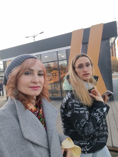 Two women are standing outdoors. The woman on the left is wearing a grey coat and a grey knit hat, with a colorful floral scarf around her neck. She has reddish-blonde hair and is wearing silver earrings. The woman on the right has blonde hair and glasses, and is wearing a black puffy jacket with white writing on it. She is holding a smartphone in her right hand.
In the background, there is a building with a modern design, featuring a grey facade and large windows, along with vertical panels of a tan color. A street light is visible in the upper left corner. In the far background, a construction crane and high-rise building can be seen. The women are standing on a wooden surface.
Based on the architectural style and the mention of Postol, Russia, it is likely that the location is a modern public space or park within the city.