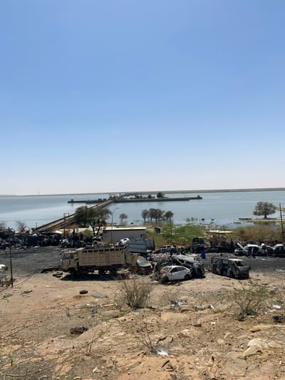 The image shows a scene of destruction in Tayba Alhasanab, Sudan, located at 15.362 latitude and 32.525 longitude. In the foreground, a dirt hill descends toward a debris field. Numerous vehicles, including a large truck, a van, and numerous cars, are charred and damaged, likely from an explosion or fire. The debris field extends towards a partially submerged dam structure, visible in the distance. Trees and sparse vegetation line the water's edge. The sky is clear and blue. There are no people visible in the image.