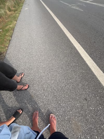The image shows a low-angle perspective of four pairs of legs and feet on an asphalt road in Prosotsani Municipal Unit, Greece. The road has a white line marking its edge and appears to be a thoroughfare. To the left of the road is a grassy verge with dry vegetation. The individuals are wearing casual footwear, including flip-flops and slides. One individual is wearing dark jeans, another is partially visible with a light blue garment. The lighting suggests it is daytime, possibly late afternoon due to the shadows. The scene captures a moment of stillness, with the focus on the feet and the immediate surroundings.