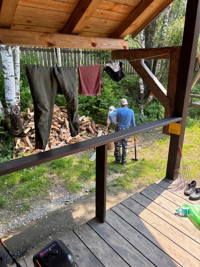 A man in a blue shirt and camouflage pants is standing on a wooden porch in Bol'shaya Rechka, Russia, holding a shovel. He is facing away from the camera and appears to be talking to another person whose back is also to the camera. Hanging on a clothesline in front of them are a pair of green pants and a red towel. Behind them, a pile of chopped firewood is stacked against a wooden fence. The porch overlooks a grassy area with trees in the background. There are also a pair of shoes, a green plastic bag, and a metal grilling basket on the porch.