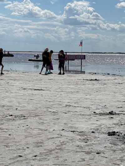 The image shows a sunny day at a salt lake. In the foreground, there is a cracked, dry salt bed with some dark rocks. In the middle ground, there is a pinkish, shallow body of water reflecting the sunlight. Several people are standing near the water's edge. One person is holding a camera. A sign with Russian text that appears to say "РОЗОВОЕ ОЗЕРО" (Pink Lake) is visible. A Russian flag is also flying on a pole behind the sign. The background shows a clear blue sky with scattered white clouds. The overall scene suggests a tourist location at a salt lake.