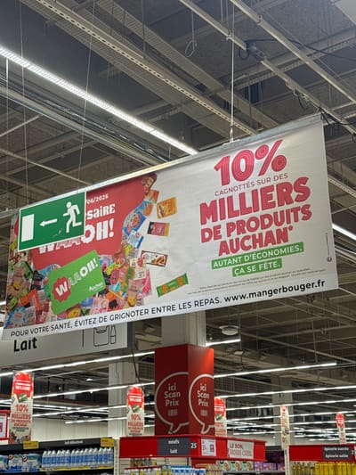 The image shows a wide-angle view of the interior of a supermarket, specifically Auchan Drap, France. A large banner hangs from the ceiling, advertising "10% CAGNOTTÉS SUR DES MILLIERS DE PRODUITS AUCHAN" (10% cashback on thousands of Auchan products). The banner features colorful illustrations of various food items and has the website "www.mangerbouger.fr" printed on it. To the left of the banner is a green exit sign with an illuminated arrow pointing left. Below the main banner, smaller signs are visible on shelves. One sign indicates "Lait" (Milk), and another has promotional material with "WAaoH!" and "10% MILLIERS DE PRODUITS AUCHAN". On the right side, there are price scanners indicated by "ici Scan Prix". Shelves in the foreground are stocked with products, including milk cartons and other grocery items. The overall environment is a brightly lit supermarket aisle.