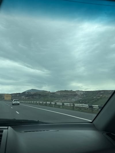 The image depicts a view from inside a vehicle, likely a car, traveling on a highway. The road surface is asphalt, marked with white lines. Adjacent to the highway is a roadside barrier, beyond which is a mountainous landscape. The sky is overcast with a layer of clouds. In the distance, a mountain peak is partially visible. There is a car traveling in the same direction on the road.
Without further evidence, it is impossible to conclusively confirm the exact location as Arzni, Armenia, though the mountainous terrain is consistent with the geography of Armenia.