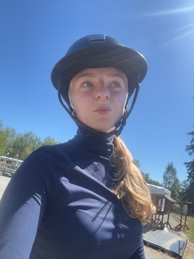A fair-skinned individual, appearing female, is centered in a close-up outdoor shot, wearing a dark blue equestrian helmet with a black brim and a dark blue long-sleeved turtleneck shirt. Her blonde hair is pulled back in a ponytail, visible from behind her right shoulder. She has light-colored eyes and her lips are pursed. A small gold hoop earring is visible on her left earlobe. She is looking upwards and slightly to the left of the frame. The background is a clear blue sky, indicating a sunny day. To the left, a multi-tiered rack holds several overturned, elongated white and light-colored boats, consistent with rowing shells or sculls. Green trees are visible behind the boats. To the right, part of a white enclosed trailer is visible, alongside a wooden structure and an overturned, reflective metallic object on the ground. The ground surface appears to be gravel or dirt. The scene is located in the Township of Langley, Canada, consistent with an equestrian or multi-sport outdoor facility.