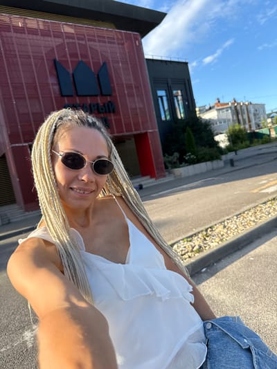 A woman with blonde braided hair and sunglasses is taking a selfie outdoors. She is wearing a white top with ruffles and blue denim. Behind her, a modern red building with a black logo is visible, along with other buildings and a street with markings. The sky is blue with some clouds. The location is Есбол ауылдық округі, Kazakhstan.