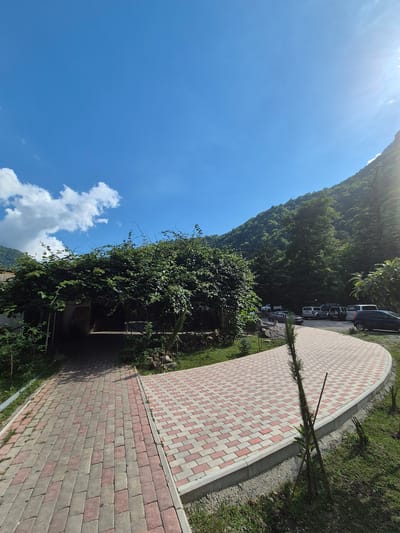 A paved walkway with a red and white checkerboard pattern curves around a grassy area in a mountainous region. Several cars are parked along the edge of the walkway, facing away from the camera, suggesting a parking area. A lush, green mountain slope covered in trees rises steeply to the left of the walkway. The sky is a bright, clear blue with a few wispy white clouds. The sun is shining brightly, casting a slight glare on the upper left portion of the image, indicating it is daytime. The overall scene is one of a scenic outdoor location, likely a tourist spot or a resting area within a natural landscape. The presence of a paved walkway and parked cars suggests some level of development in the area. There is no visible text in the image.