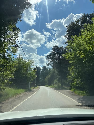 The image captures a view from a vehicle traveling on a paved road through a wooded area in Подмосковье Дпк, Russia. The road curves gently to the right in the distance. On either side of the road, dense green foliage of trees and undergrowth is visible, with some taller coniferous trees also present. The sky above is a bright blue, dotted with fluffy white cumulus clouds. Sunlight streams down through the clouds, creating lens flare effects and illuminating the scene. Power lines are strung across the sky, supported by poles visible along the side of the road. The foreground of the image shows the hood and windshield of the vehicle from which the photo was taken. The overall impression is that of a scenic drive through a natural landscape on a sunny day.