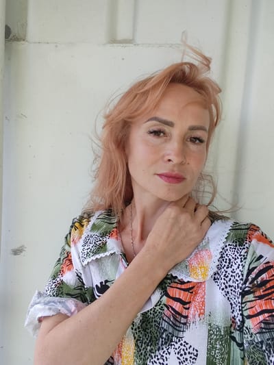 A medium shot captures a woman with light reddish-blonde hair and fair skin, posing against a neutral, off-white background. She is looking directly at the camera with a slight smile. Her hand is raised, with her fingers resting gently on her neck. She has dark eyebrows and dark, winged eyeliner, and her lips are painted a muted pink. She is wearing a colorful shirt with a white collar trimmed with white lace. The shirt features a bold, abstract pattern with various colors, including green, orange, yellow, black, and white, with some sections resembling animal prints. Her hair is windswept, with strands blowing across her forehead and to the side. The lighting suggests it is daytime, possibly outdoors given the wind. The background appears to be a textured, possibly corrugated surface, with some subtle marks and imperfections, hinting at an industrial or utilitarian setting. There is no visible text in the image.