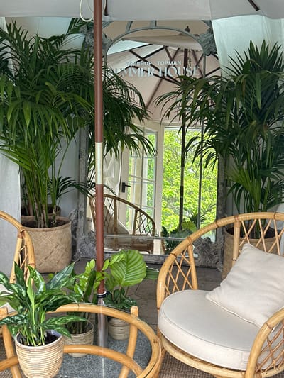 The image depicts an outdoor seating area, likely a patio or covered terrace, with lush green tropical plants. In the foreground, there are two rattan chairs, one with a light-colored cushion. A circular glass-topped coffee table sits between them, holding several potted plants. A large umbrella pole is visible in the center of the frame, and a large ornate mirror is positioned behind the plants, reflecting a doorway and trees outside. The text "TOPSHOP TOPMAN SUMMER HOUSE" is visible in the reflection. The overall ambiance is tropical and relaxed. This scene is located at The Draper's Yard, 38 North Street, Chichester, West Sussex, PO19 1LU, United Kingdom.