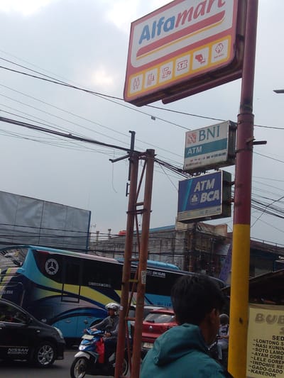 The image captures a street scene in Megamendung, Indonesia, under an overcast sky. A prominent red and white Alfamart sign hangs overhead, displaying various icons. Below it, two ATM signs are visible, one for BNI and another for BCA. A large blue and yellow bus with the "JETBUS PANSATYA" branding is parked on the left, with "BLUE STAR" written on its side. A black Nissan car is also present, and a person on a scooter, wearing a helmet, is riding past. In the foreground, the back of a person wearing a teal hoodie is visible. The environment appears to be urban, with power lines crisscrossing the sky and buildings in the background. A yellow pole with some text and an orange object on top is also in the frame.