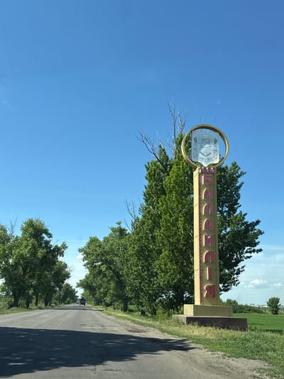 The image shows a large, retro-style roadside sign under a clear, bright blue sky on a sunny day. The scene depicts a rural or semi-rural environment, with a dirt or gravel road on the right, bordered by green grass and numerous lush green trees. In the distance, an open field and faint structures are visible beneath a light blue sky with a few clouds.
The prominent sign features a tall, yellowish rectangular base with bold red Cyrillic letters. The visible text on the sign spells out "БАЛАКЛЕЯ" (Balakliya), indicating the location. At the top of the sign, encased in a golden circular frame, is a white emblem. This emblem contains a stylized drawing that appears to include agricultural motifs such as wheat stalks, possibly a sheaf, and other indistinct elements, resembling a local crest or coat of arms. No people are present, and no specific activity is occurring beyond the static display of the sign in its natural setting. The time of day appears to be midday or early afternoon due to the bright lighting.