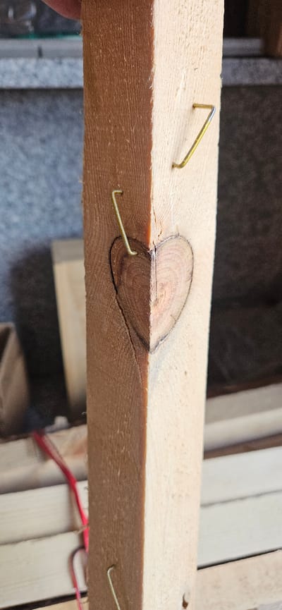 The image shows a close-up, low-angle shot of a wooden plank with several staples embedded in it. The plank has a visible wood grain and a knot. One staple is clearly visible in the foreground, angled upwards from the wood. Another staple is partially embedded near the knot. In the background, more wooden planks are visible, along with some red wires, suggesting a possible construction or repair context. The lighting appears to be artificial, casting soft shadows and highlighting the texture of the wood. There is no visible text in the image. The scene is set outdoors, possibly in a workshop or construction site, given the materials and tools visible. There are no people or animals in the frame. The focus is entirely on the wooden plank and the staples.