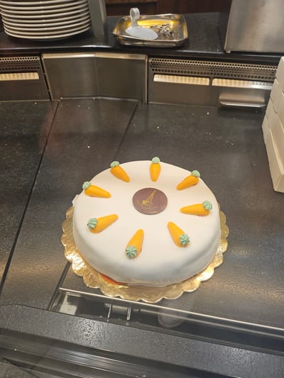 A white frosted cake, decorated with nine small orange carrot-shaped candies with green frosting tops, sits on a gold scalloped cake board on a black countertop. The cake is round and has a dark brown circular emblem in the center, depicting a crane and text that appears to read "Konditorei Sprüngli Zürich". The countertop is speckled and appears to be in a commercial setting, possibly a bakery or cafe, with stacked plates visible in the background to the left and a stainless steel apparatus to the right. The overall impression is of a well-presented dessert item. The location of this scene is the Konditorei Sprüngli in St. Moritz, Switzerland, a renowned confectionery shop established in 1859.