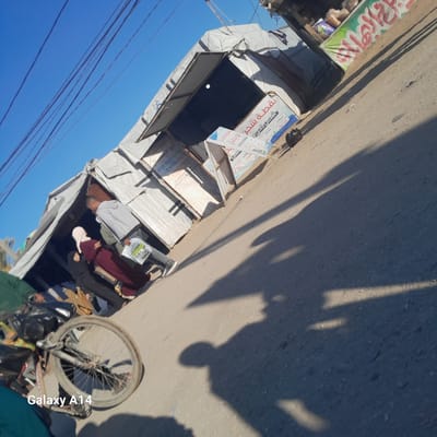 The image, taken in what appears to be Al-Mawasi, Unknown, shows an outdoor street scene under a clear blue sky. A bicycle with a flat front tire leans against a structure to the left. Several individuals are near what appears to be a makeshift store or market stall covered with canvas. One woman wears a maroon dress and a light pink hijab, while a man next to her is dressed in grey. The store's signage indicates services like "mobile charging" (نقطة شحن) and possibly "phone recharge" (رصيد جوال) in Arabic. The ground in the foreground is marked by long shadows, and electrical lines are visible in the upper right corner.