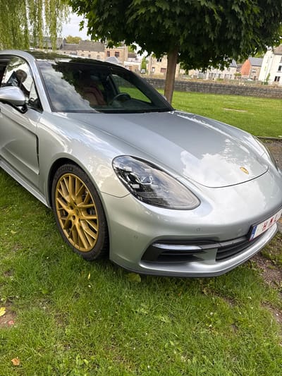 A silver Porsche Panamera with gold rims is parked on a grass lawn next to a tree in Melreux, Belgium. The front of the car is facing right and slightly up. The license plate is visible but partially obscured. Buildings are visible in the background through the car windows and beside the tree, indicating an urban or semi-urban setting. The scene appears to be outdoors during daylight.