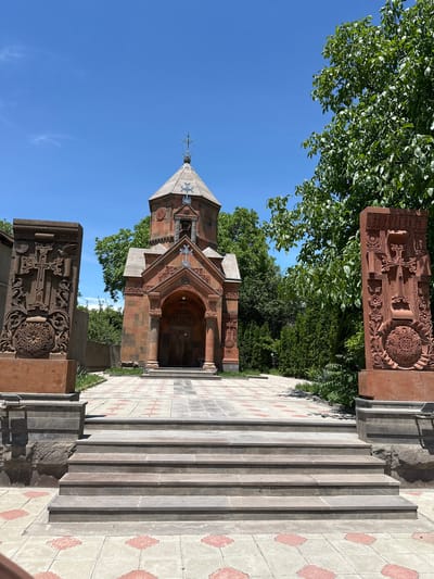 The image presents an outdoor scene in Garni, Armenia, featuring an Armenian Apostolic church under a clear blue sky. A central reddish-brown stone church with an octagonal drum and conical roof, topped by an ornate metal cross, is the primary subject. Its facade includes an arched portico detailed with intricate stone carvings and integrated crosses. In the foreground, a five-step stone staircase leads to a paved pathway composed of light-colored tiles with reddish-orange geometric patterns. Flanking this pathway are two tall, rectangular, reddish-brown stone stelae (khachkars), each meticulously carved with Armenian crosses, stylized birds, and complex floral-geometric designs. The church and pathway are enveloped by lush green foliage, including broadleaf trees and dense coniferous shrubs. The sky is bright blue with minimal, wispy clouds. No individuals are visible.