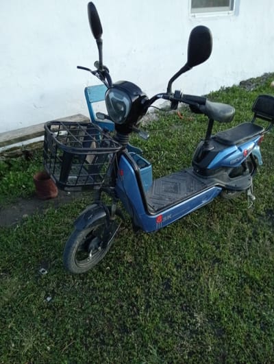 An outdoor medium shot captures a blue electric scooter parked on green grass in front of a white building. The scooter is positioned at a slight angle, facing towards the right. It has a black front basket, a black seat, and black handlebars with mirrors. A blue seatback is visible behind the main seat. The scooter has the branding "GIDIAN" and "SK" on its side. To the left of the scooter, a rusty bucket is placed on a dirt path. The white building behind the scooter has a horizontal window with a light-colored frame. The ground is covered in green grass with some patches of dirt. The image appears to have been taken during daylight.