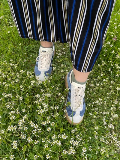 An overhead shot shows a person standing in a field of green grass and tiny white flowers. The person is wearing striped pants with black, white, and blue stripes and light-colored Adidas sneakers with beige soles. Green socks are visible between the pants and shoes. The location is not specified in the image, so it is not possible to determine if it is in Barkach, Bulgaria.