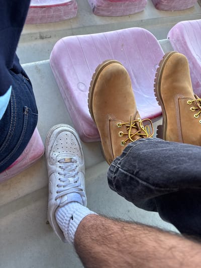 The image shows a ground-level shot of two people sitting in a stadium. The person on the left is wearing dark blue jeans and Timberland boots. The person on the right is wearing black jeans and a white Nike Air Force 1 shoe with a white sock. They are sitting on light purple stadium seats. The location is presumed to be in the city of Valpaços, Portugal.