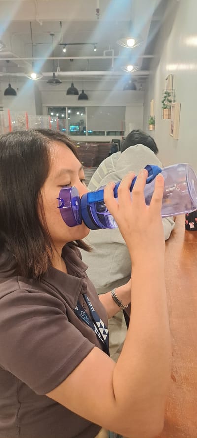 A woman is holding a blue water bottle and drinking from it. She is wearing a gray shirt and a lanyard with some text on it. The scene appears to be indoors, possibly in a cafe or restaurant, with lights and some decorative elements visible in the background. The woman's expression suggests she is focused on drinking. The orientation of the image is sideways. There is text on the lanyard which is difficult to read due to the image quality and angle.