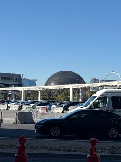 The image depicts a daytime scene in Las Vegas, Nevada, United States of America, showing a street-level perspective with prominent architectural landmarks in the background.

In the foreground, the lower portion of a red-orange plastic traffic barrier is visible on the left, next to a rocky, unpaved strip of ground. An asphalt road occupies the immediate mid-ground. Concrete barriers and metal fencing separate the road from a large outdoor parking lot filled with numerous parked vehicles, primarily dark-colored sedans and SUVs. A red-on-white "NO PARKING ANY TIME" sign is affixed to the metal fence. A black four-door sedan (possibly a Toyota Camry) is prominently positioned on the asphalt road, facing right, with its front half visible. Behind and partially to the right of the black sedan, the front portion of a white panel van is visible.

Above the parking lot and vehicles, an elevated white concrete monorail track, supported by multiple columns, spans horizontally across the frame. Green foliage, consisting of trees and bushes, is visible beneath and behind the monorail structure.

In the background, dominating the scene, is the large, dark, spherical structure of the Sphere at The Venetian Resort. To its left, a modern, multi-story building with a flat roof is partially visible. To the far right, beyond the Sphere, the upper arc of a large white Ferris wheel, identified as the High Roller, is visible against the clear blue sky. Other urban buildings are visible in the distance. The sky is bright and cloudless, indicating clear weather. No individuals are distinctly visible or engaged in specific actions.