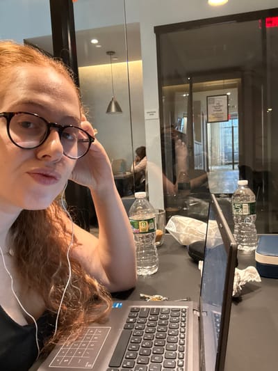 A female individual is depicted in a close-up selfie in an indoor setting within Plainsboro Township, United States. She possesses long, wavy, reddish-brown hair, wears dark-rimmed eyeglasses, and has white wired earbuds inserted. Her left hand is raised, touching her temple, with a ring visible on her left ring finger. Her lips are pursed. She is dressed in a dark top. On the dark table in front of her are an open gray laptop with an angled screen, a set of silver keys, two plastic Poland Spring water bottles, a glass with brown liquid, a crumpled white tissue, a dark eyeglasses case, and a small blue rectangular object. The background features glass partitions indicative of a communal or office area. Through these, another individual is visible seated at a table. A silver pendant light fixture hangs from the ceiling. A reflective surface displays parts of the foreground subject's arm. Beyond, a lit corridor is visible, suggesting an exterior window view.