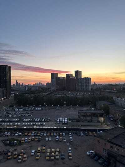 This high-angle urban landscape image, captured at dusk or sunset, provides a sweeping view of a city from above. The sky dominates the upper portion, transitioning from a soft grey-blue on the left to vibrant hues of orange, yellow, and red near the horizon on the right. A prominent, elongated streak of pinkish-red clouds stretches across the middle of the sky, catching the last light of day.
In the mid-ground, several tall, modern buildings, likely residential or commercial skyscrapers, rise against the colorful sky, their facades featuring numerous windows. Further in the distance, a sprawling city skyline recedes into the twilight.
The foreground and mid-ground below the buildings are characterized by extensive, somewhat gritty urban infrastructure. Multiple large parking lots, paved with asphalt or dirt, are filled with numerous vehicles. A striking feature is the high concentration of cars that appear to be taxis, identifiable by their yellow, white-and-yellow, or dark colors, parked in neat rows and scattered arrangements.
Interspersed among the parking areas and buildings are a few clusters of trees. Older, darker industrial-looking buildings and warehouses with distinct rooflines are also visible, hinting at a mix of old and new urban development. Streetlights are beginning to illuminate, casting pools of light onto the ground and suggesting the transition from day to night. No specific human activity is discernable, but the parked vehicles imply a bustling urban environment. No visible text can be clearly read. The scene depicts an urban area, consistent with the provided location context of Ukromne, Ukraine.
