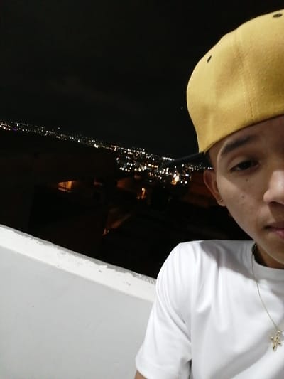 A young person wearing a yellow baseball cap and a white T-shirt poses for a selfie. They are wearing a gold cross necklace. The background is a cityscape at night, with many scattered lights across a dark horizon. A short white wall appears in the foreground, suggesting the person is on a balcony or elevated platform. The image has low lighting and a slight upward angle.