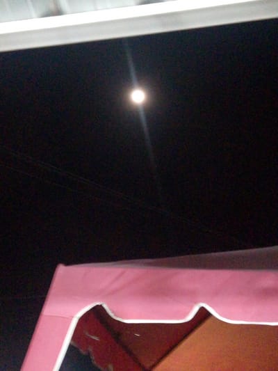 The image shows a nighttime scene with a bright full moon illuminating a dark sky. A thin band of a white structure, possibly a roof or awning, is visible at the top of the frame. Below the moon, two thin lines are faintly visible, which might be power lines or cables. In the foreground, the top portion of a pink tent or canopy with a white trim is seen, casting a shadow. The overall impression is of an outdoor setting at night, possibly in a rural or less urbanized area given the clear view of the moon and the presence of the tent. The mood is peaceful and serene, highlighted by the bright moonlight against the dark sky. There is no visible text in the image.