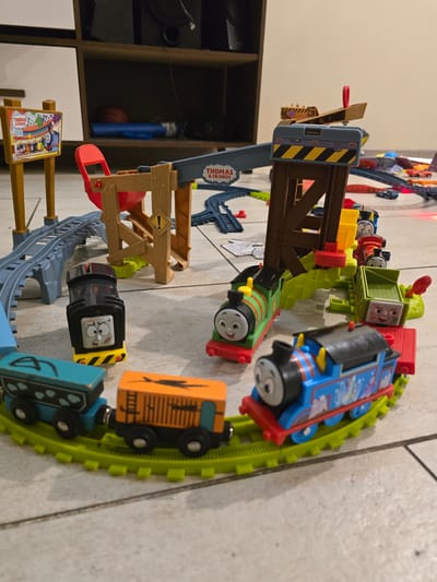 The image shows a Thomas the Tank Engine toy train set on a tiled floor. The set includes various trains such as Thomas, Percy, and other character trains, along with tracks, a crane, and other accessories. The train set is arranged in a circular and branching configuration. A sign post is visible at the bottom. In the background, a brown cabinet with shelving is partially visible, containing a blue bottle and a red ball. The scene is indoors. The location in the city of La Florida, Chile is not determinable from the image.