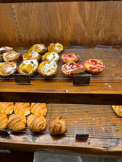 The image shows a bakery display case in Arakawa City, Japan (35.728, 139.766). The case has two levels and is made of wood and metal. The upper level has several pastries, including two with fruit and a few with powdered sugar. The lower level also has several pastries, all of which are plain and have a flaky, buttery texture. All of the pastries are on a metal grid. There are small price tags on the display case.  There are no people in the image.  There is no interaction taking place in the image. The environment is a bakery.