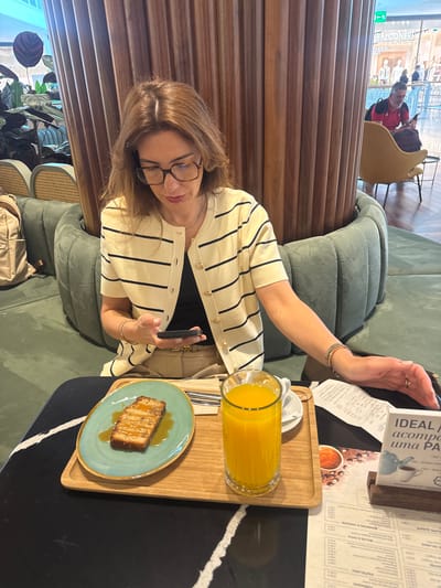 In a casual café setting in Senhora da Hora, Portugal, a woman is seated at a table, enjoying a snack and drink. She has shoulder-length brown hair and wears glasses, a cream-colored cardigan with black stripes, and a black top. Her left arm is extended towards a menu stand, while her right hand holds a dark smartphone.
On the dark-colored table with white accents, a wooden tray holds a slice of cake or bread with caramel drizzle on a light blue plate, and a tall glass of orange juice. A paper receipt is visible near the menu stand. The woman is seated on a green upholstered bench, with a light-colored bag resting on the floor beside her. In the background, wooden slatted partitions provide a dividing element, and reflections reveal other patrons, including a person in a red shirt, and furniture like a yellow armchair and rattan-style seating. Green plants add to the decor.
Visible text from the menu includes "PASTELARIA," "BOLOS A FATIA" (cakes by the slice), "BOMBONS À UNIDADE" (chocolates by the unit), and phrases like "IDEAL acompanha uma PA" (IDEAL to accompany a pastry). Prices such as "1.00€" and other numbers are also legible. The word "FALCONERE" can be seen in the background reflection. The atmosphere appears relaxed, suggesting a coffee break or casual meal.