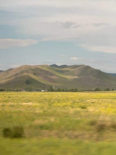 The image depicts a vast, undulating landscape under a pale sky with scattered clouds. In the foreground, a blurred field of green and yellow suggests motion, possibly from a vehicle. In the mid-ground, a line of trees separates the field from rolling hills covered in green vegetation. A small white building and a tall, red and white striped industrial smokestack are visible among the trees. In the background, distant mountains stretch across the horizon. The scene is likely a rural or semi-rural area given the natural landscape and the presence of industrial elements. The location of Ust'-Kamyshta, Russia, is provided as context for this image.