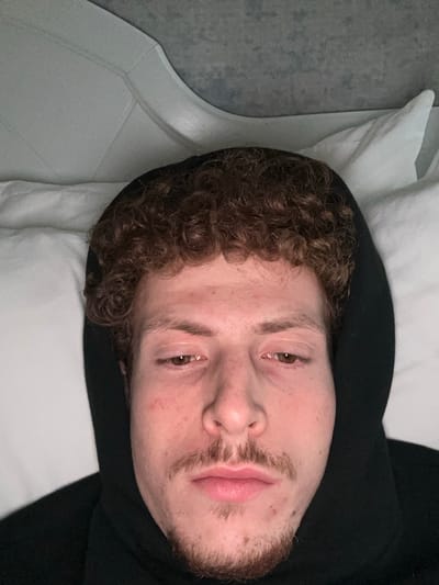 A man with curly brown hair and a patchy beard rests in bed. He is wearing a black hooded sweatshirt. His eyes are open. He appears to be lying on a white pillow on a white bed, with a light gray wall or surface partially visible in the background to the left. He is positioned at an angle to the camera, with his head tilted to the right.