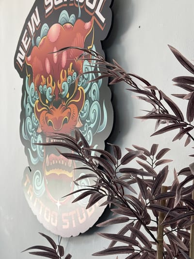 This media file captures a close-up view of a tattoo studio sign, partially obscured by artificial bamboo-like branches with dark brown leaves. The sign itself is shaped with decorative scalloped edges and features a colorful illustration. Dominating the center of the sign is a stylized representation of a mythical creature, possibly a lion or dragon, depicted in shades of red and orange with yellow accents. Swirling patterns resembling clouds or stylized waves in teal and blue surround the creature's head. Visible text on the sign includes "NEW" in white block letters on the left side, and "TATTOO STUDIO" in orange letters curving around the right side. The background is a plain, textured white surface, likely a wall or ceiling. The lighting suggests an indoor environment, possibly during the day, with a shadow cast on the white surface from the sign and branches, indicating a light source from above and to the left. The overall impression is one of a vibrant and perhaps slightly edgy establishment, with the artificial foliage adding a touch of decorative flair. The location context of Campillo Del Moro, Spain, does not provide specific visual cues within the image itself.