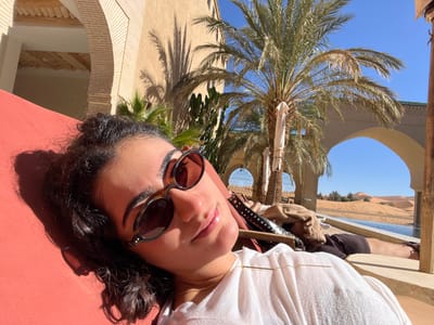 The scene depicts two individuals relaxing outdoors at a resort facility in Rissani, Morocco, bordering a desert landscape. In the immediate foreground, a female subject with dark, curly hair, wearing round tortoiseshell-patterned sunglasses and a white top with a gold bar necklace, lies on her back on an orange-red sun lounger, facing the camera. To the right, partially visible, a second individual lies on a dark-colored sun lounger adjacent to an infinity-edge swimming pool. The pool, filled with blue water, extends towards extensive golden sand dunes of the Sahara desert, visible under a clear blue sky. Architectural elements include light-colored, traditional Moroccan structures with multiple arches, one of which frames the desert view. Several tall palm trees are strategically placed, casting distinct shadows across the sun-drenched scene. Sparse green vegetation is present near the architectural structures and on some distant dunes.