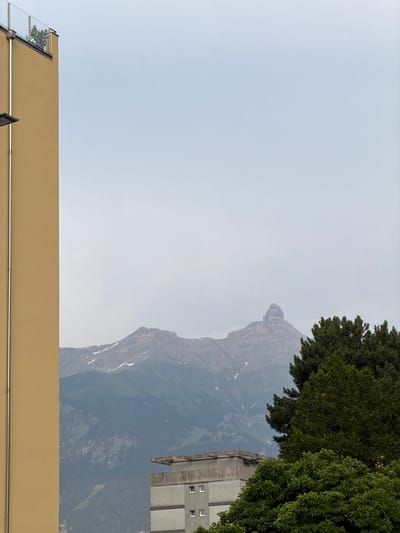 The image displays a view of a dramatic mountain peak in the background, likely in Monthey, Switzerland. The mountain is imposing, with steep, rocky slopes and patches of snow still visible, suggesting a cooler season or high altitude. A dark green, dense forest covers the lower slopes of the mountain. In the mid-ground, a modern building with a concrete facade and several windows is visible. The building appears to be several stories high. In the foreground, a tan-colored wall runs horizontally across the bottom of the image, possibly part of another building or structure. A section of a glass railing is visible on the left side of the tan wall. The sky is a pale, hazy blue, suggesting overcast conditions or a distant haze, possibly indicating a time of day without direct sunlight, or a generally hazy atmosphere. There are no people or clear signs of activity, giving the scene a still, natural feel. The composition is unusual as the image is oriented sideways.
